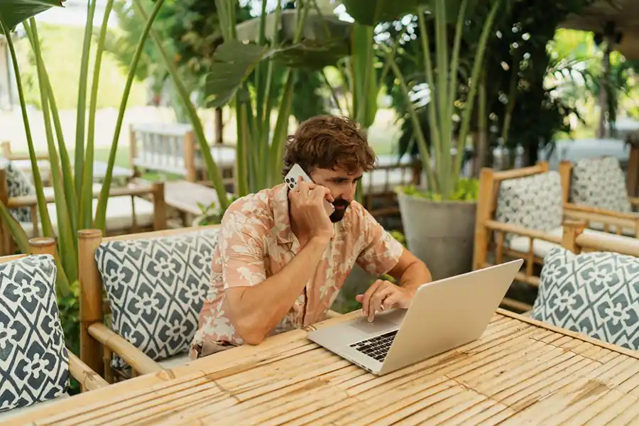 Handsome man with beard using lap top and mobyle phone sitting in outdoor cafe with tropical interior